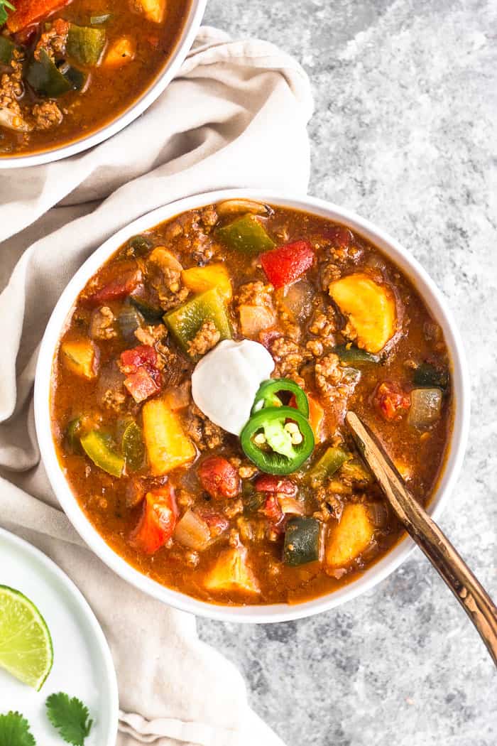 White bowl filled with Whole30 beef and plantain chili topped with coconut yogurt and sliced jalapeños. A gold spoon is sticking out of the bowl. Around the bowl is a tan linen, another bowl of chili, and a plate with lime wedges.