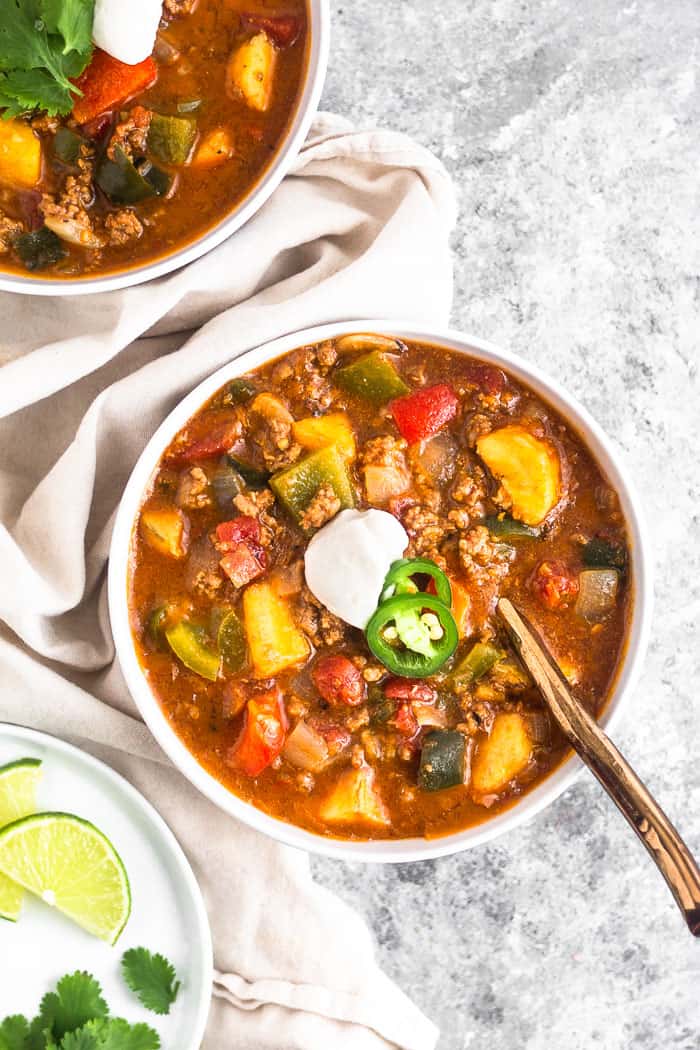 White bowl filled with paleo beef and plantain chili topped with coconut yogurt and sliced jalapeños. A gold spoon is sticking out of the bowl. Around the bowl is a tan linen, another bowl of chili, and a plate with lime wedges and cilantro.