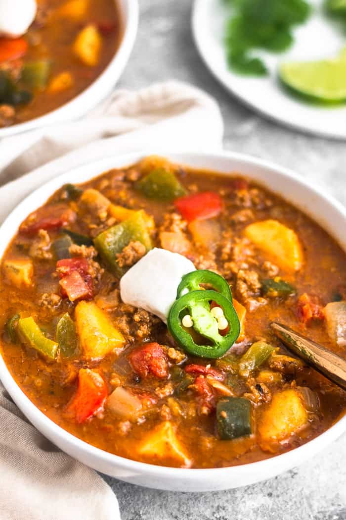 Close up a large bowl filled with beef and plantain chili topped with yogurt and sliced jalapeños. A spoon is in the bowl and behind the bowl is another bowl and a plate filled with lime wedges and cilantro.