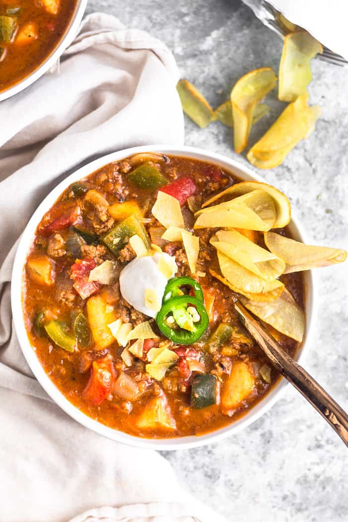White bowl filled with beef and plantain chili topped with coconut yogurt. sliced jalapeños, and chips. Next to it is more chips, another bowl of chili, and a tan linen.