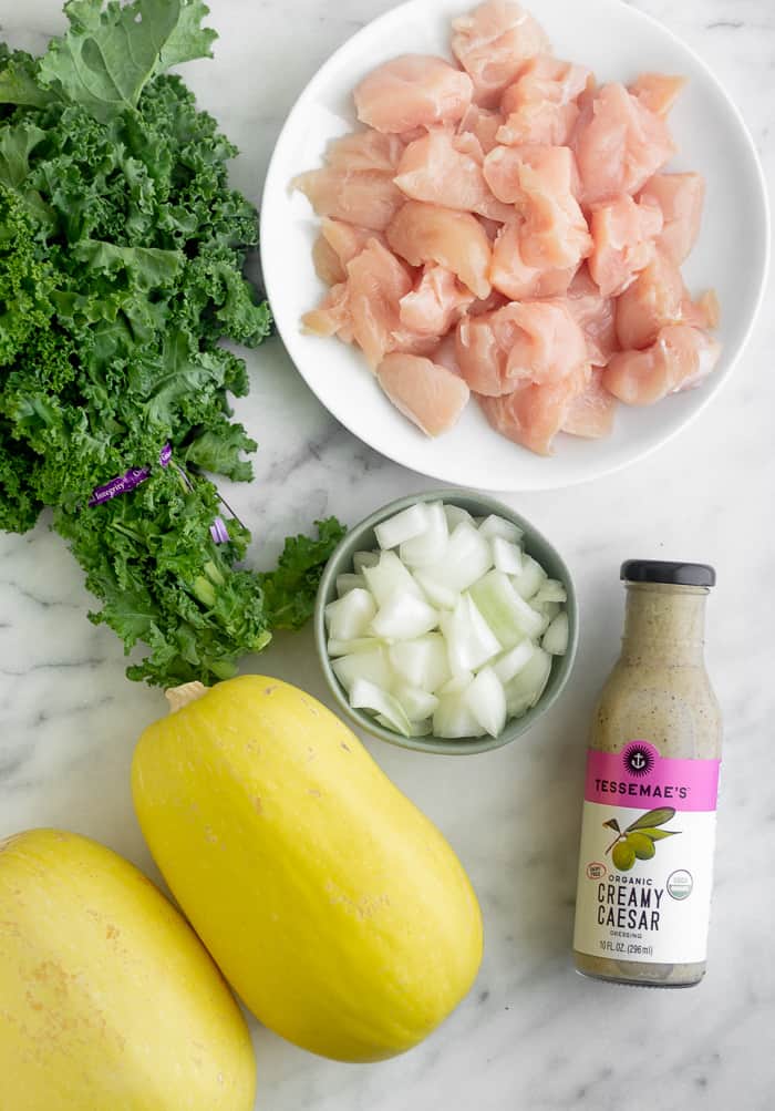 A bunch of kale, plate of rase cubed chicken, bottle of caesar dressing, bowl of chopped onions, and 2 spaghetti squash sitting on a counter.