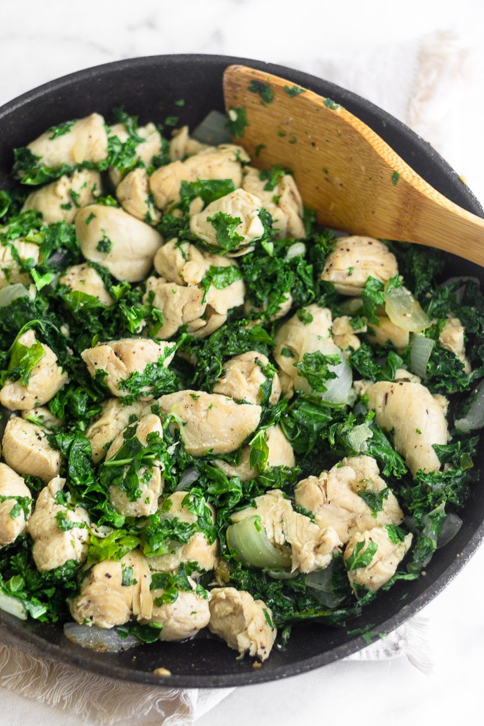 Large saute pan filled with cooked chicken, onions, and kale. A wooden spoon is sticking out of it.