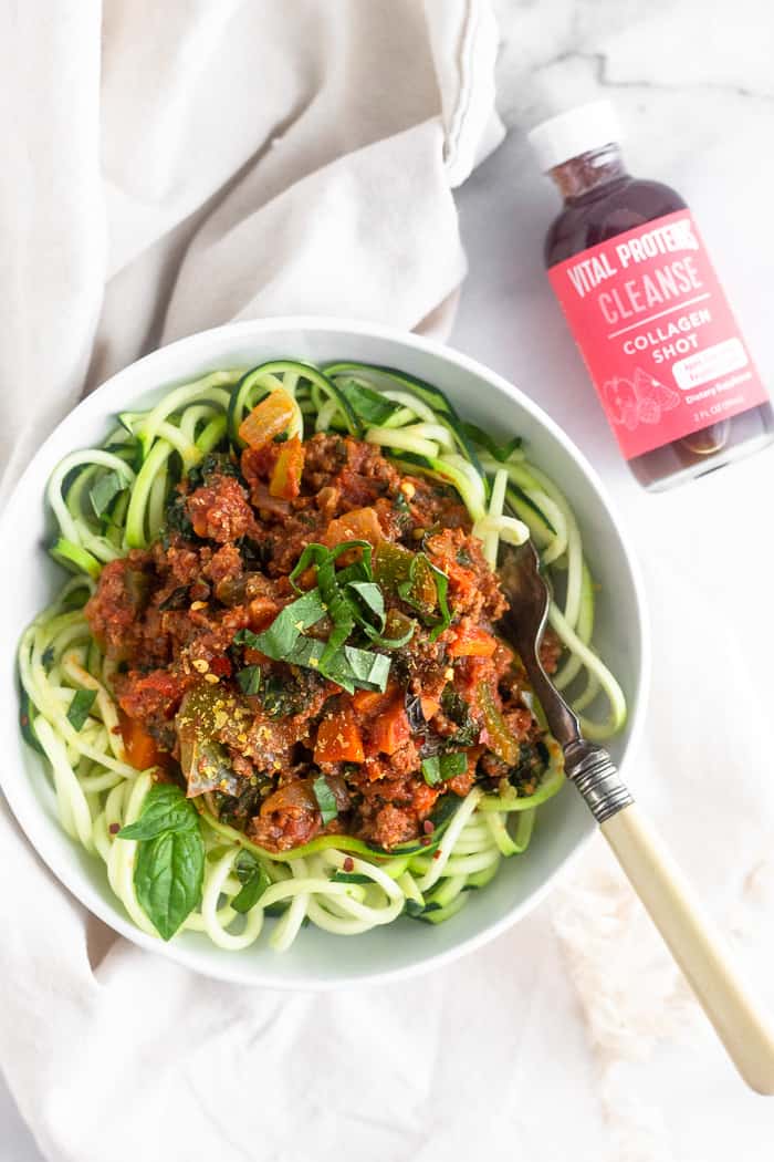 A bowl of zucchini noodles and meat sauce in a white bowl with a fork resting in the bowl. Next to is a tan linen and a cleanse collagen shot.