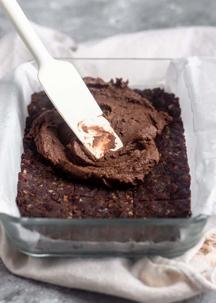 Glass dish filled with raw walnut protein brownies and topped with chocolate protein frosting on top and a spatula spreading it out.
