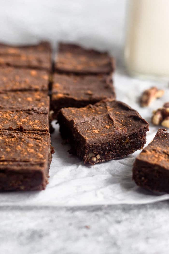 Raw no bake walnut protein brownies lined up as if they were just taken out of the pan. The front right is off to the side and almost out of the picture while the second one is just slid out a little. Around them are some walnuts and a jar of milk.