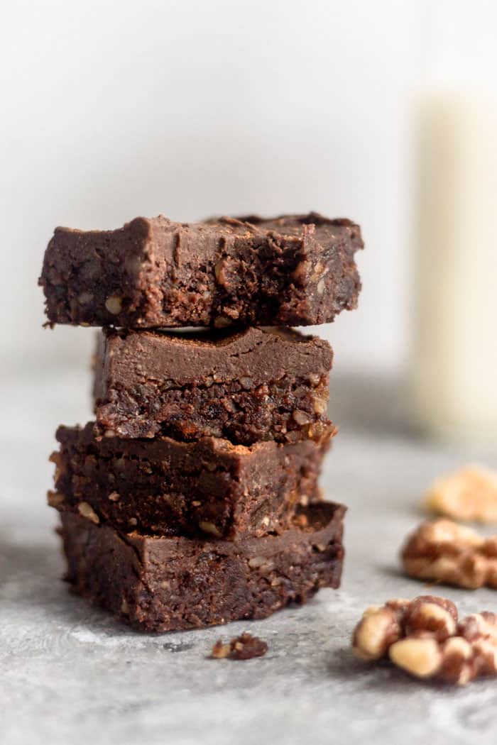 A stack of 4 vegan no bake walnut protein brownies with the top one having a bite taken out of it. They are surrounded by a few walnuts and behind them is a jar of milk.