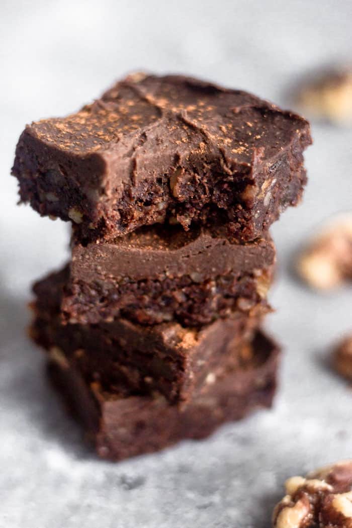 A stack of 4 vegan no bake walnut protein brownies with the top one having a bite taken out of it. They are surrounded by a few walnuts.