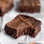 A vegan no bake walnut protein brownie with a bite out of it sitting on a small pieces of white paper. Behind it is more brownies and in front of it is a walnut.