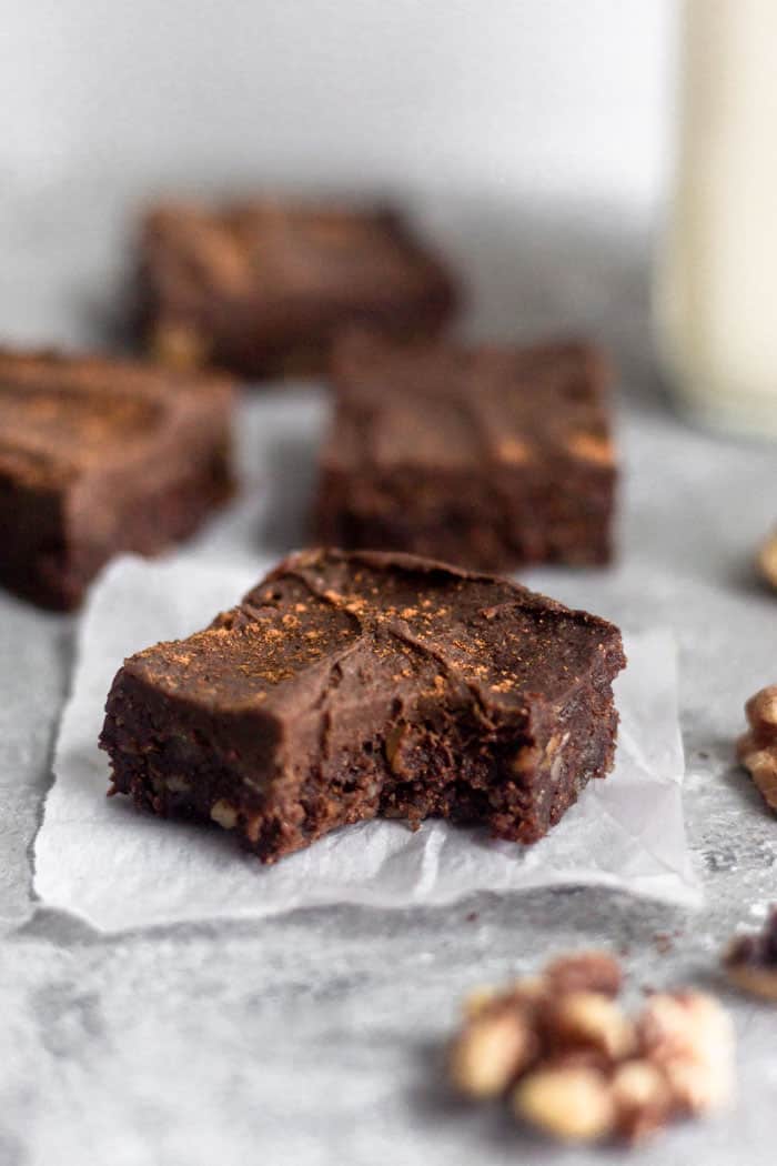 A raw no bake walnut protein brownie with a bite out of it sitting on a small pieces of white paper. Behind it is more brownies and a jar of milk.