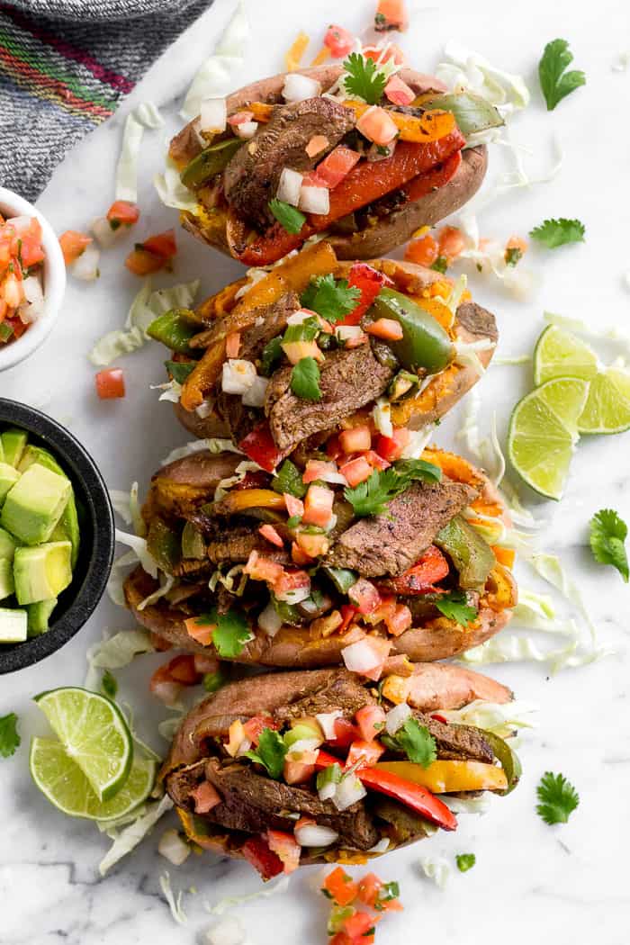 Overhead shot of a line of fajita stuffed sweet potatoes topped with pico de Gallo. Next to them is a bowl of cubed avocado, pico de Gallo, and lime wedges.