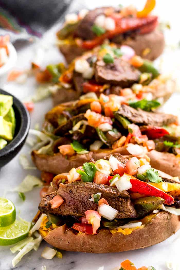 Steak fajita stuffed sweet potatoes stopped with pico de Gallo next to a bowl of cubed avocado and lime wedges.