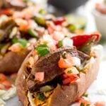 A close up of steak fajita stuffed sweet potatoes topped with pico de Gallo. Behind them is a bowl of salsa and cubed avocado.