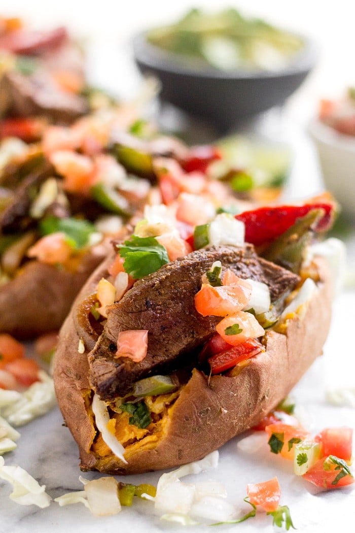 A close up of steak fajita stuffed sweet potatoes topped with pico de Gallo. Behind them is a bowl of salsa and cubed avocado.