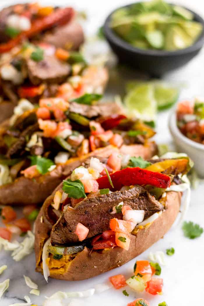 Steak fajita stuffed sweet potatoes topped with pico de Gallo and cilantro. Behind them is a bowl of salsa, lime wedges, and a bowl cubed avocado.