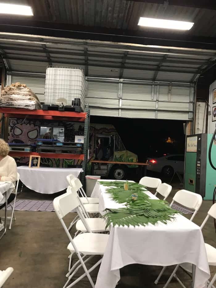 Back of a brewery with tables set up with palms on top of them. A large garage door is open with a door truck in the back.