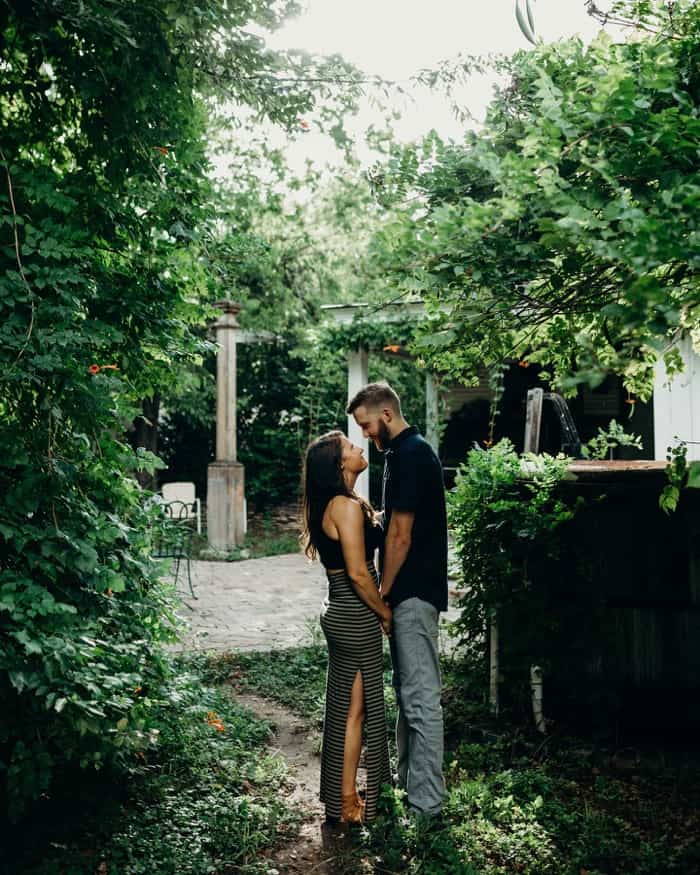 An engaged couple standing in some greenery holding hands and embracing each other.