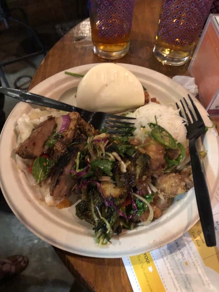 Large paper plate filled with rice, Brussel sprouts, chicken, pork, and a bun.