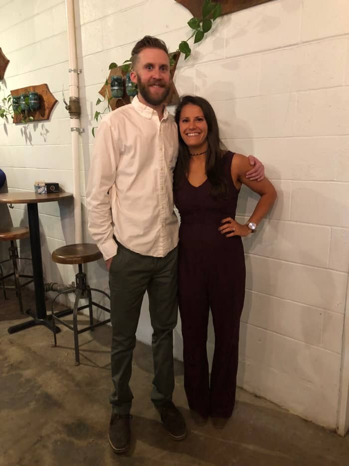 Couple at their rehearsal dinner posing for a picture. Girl has dark long hair and is wearing a jumpsuit. The guy has short brown hair and wearing a white collar shirt.