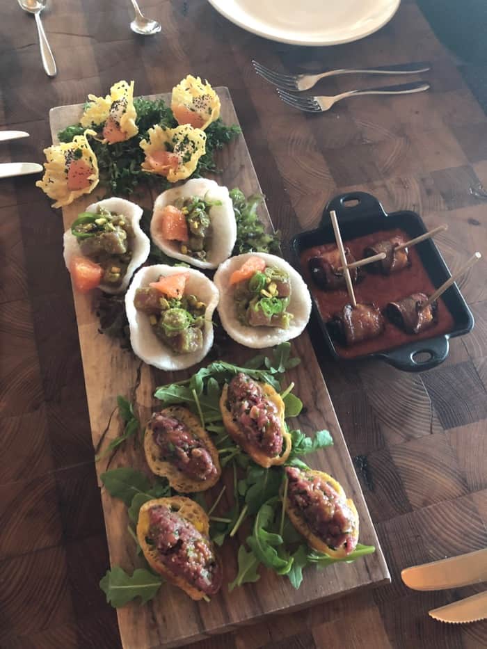 Wooden serving board filled with 3 different appetizers. Next to it is a small cast iron filled with sauce and bacon wrapped dates.