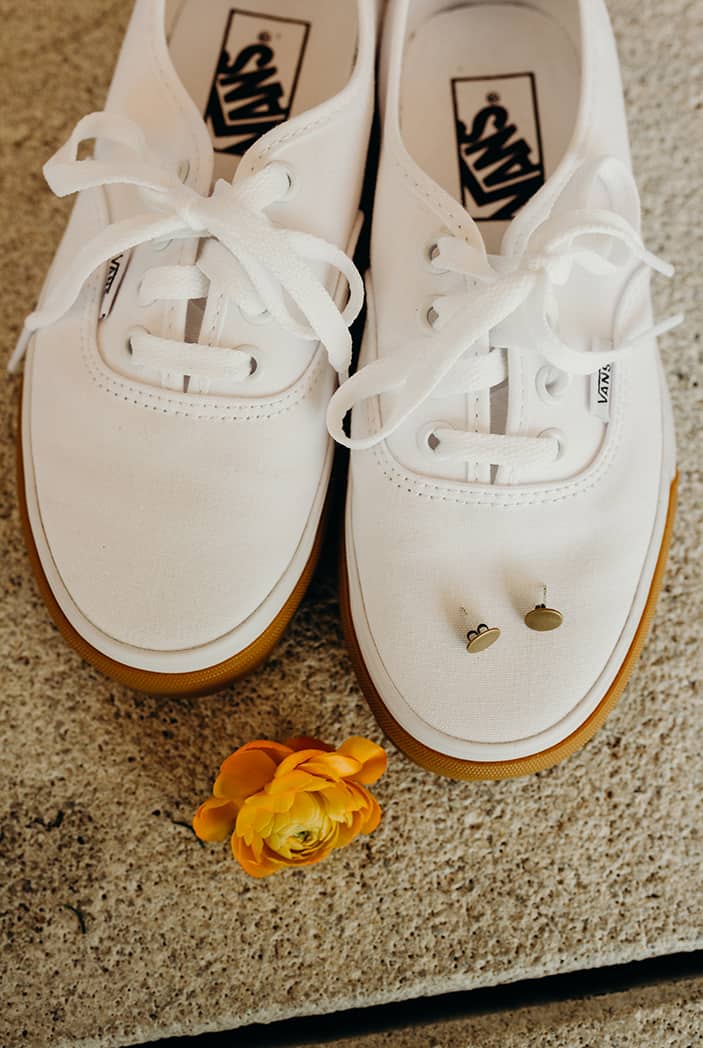 White vans with gold studs sitting on top of them. Next to them is an orange flower.