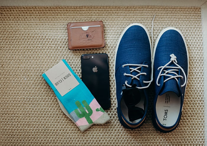 Men's blue TOMs, a pair of socks with a cactus on it, an iPhone, and a leather wallet.