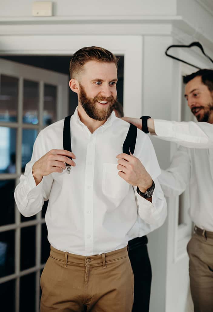 A groom getting ready for his wedding with his friend helping put on his suspenders.