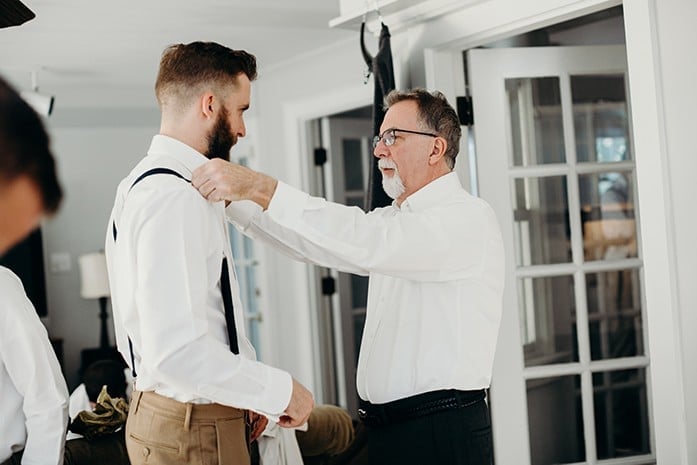 A son getting ready for his wedding with his dad helping put on his suspenders.