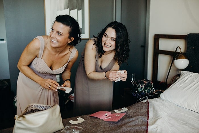 Two bridesmaids opening presents in an hotel room looking off to the side.