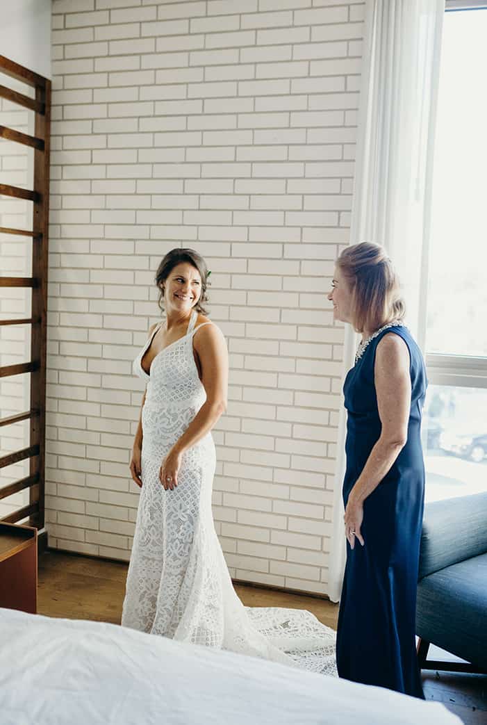 A bride in her dress turning to see her mom for the first time while getting ready in a hotel room.