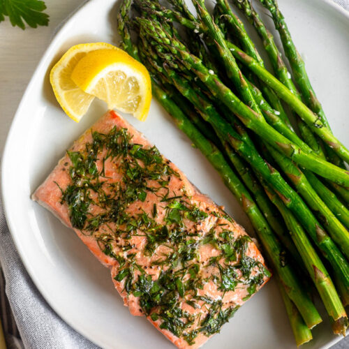 Dill coated salmon and asparagus on a white plate with two lemon wedges next too it. It is on a grey towel with fresh parsley next to it.