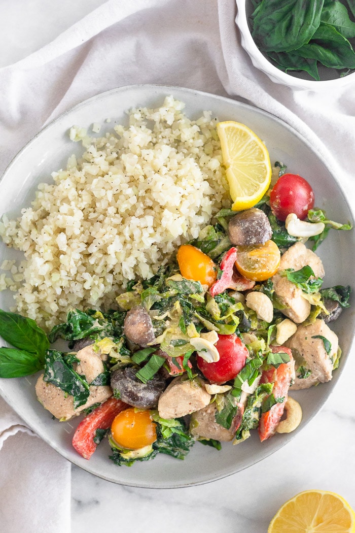 White plate filled with one pan creamy balsamic chicken and veggies, cauliflower rice, fresh basil, and a lemon wedge.