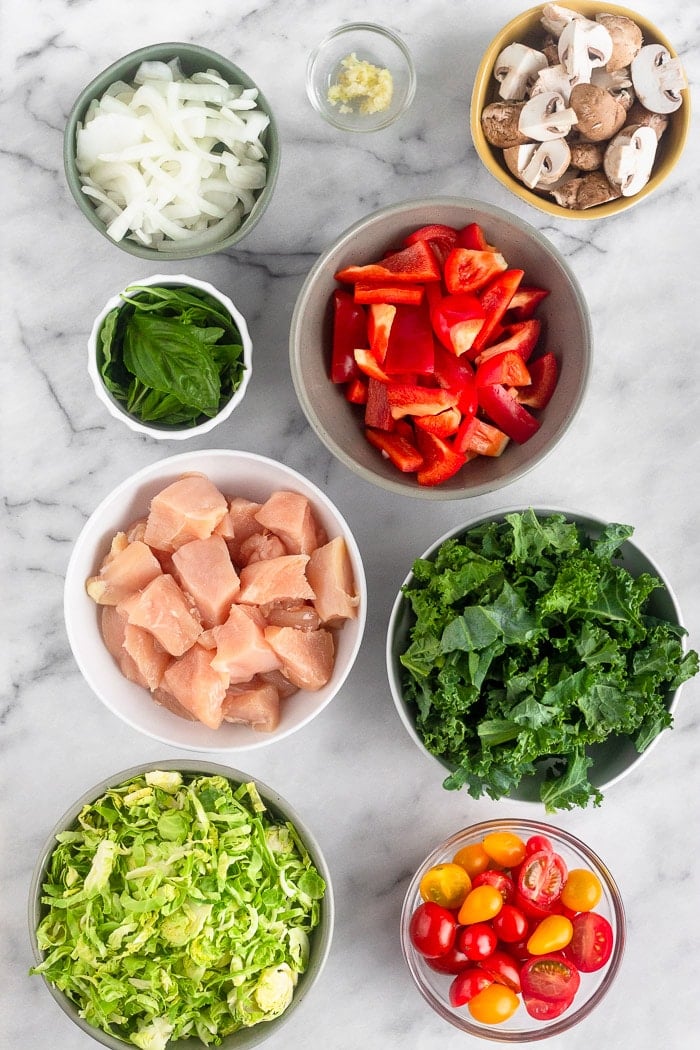 White marble counter top filled with the ingredients to make balsamic chicken and veggies skillet. There is onion, garlic, mushrooms, peppers, kale, tomatoes, Brussels sprouts, chicken, and basil.