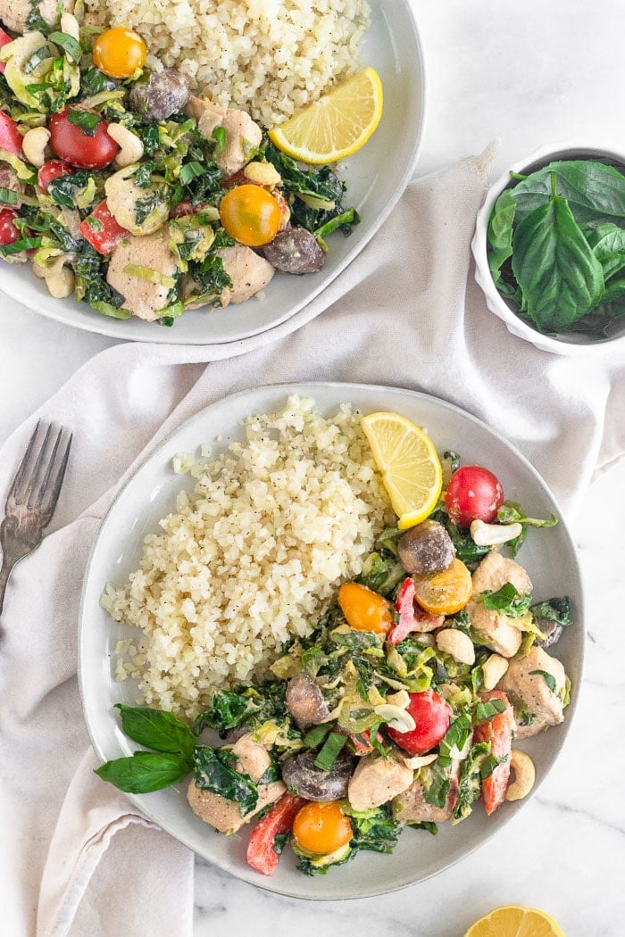 Overhead of two plates filled with one pan creamy balsamic chicken and veggies and cauliflower rice topped with fresh basil and a Leon wedge. A bowl of basil is next to them.
