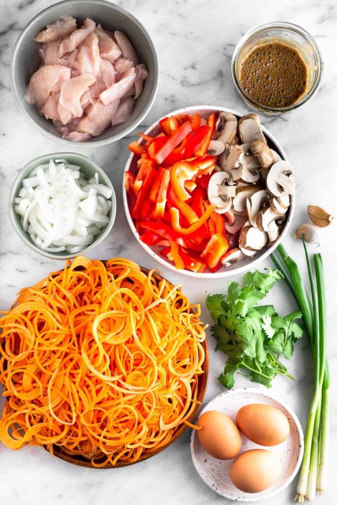 Marble counter with a jar of pad thai sauce, a bowl of sliced bell peppers and mushrooms, 2 garlic cloves, 3 stalks of green onions, a bunch of cilantro, a small plate with 3 brown eggs on it, a bowl of sweet potato noodles, a bowl of sliced onions, and a bowl of sliced raw chicken.