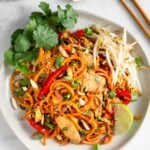 White plate with chicken pad thai made with sweet potato noodles topped with bean sprouts, cilantro, and a lime wedge. Next to it is chopsticks, lime wedges, and cilantro.