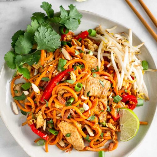 White plate with chicken pad thai made with sweet potato noodles topped with bean sprouts, cilantro, and a lime wedge. Next to it is chopsticks, lime wedges, and cilantro.