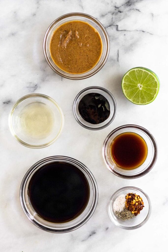 Marble counter with a bowl of almond butter, half a lime wedge, a bowl of tamarind paste, a bowl of fish sauce, a bowl of spices, a bowl of coconut aminos, and a bowl of rice vinegar.