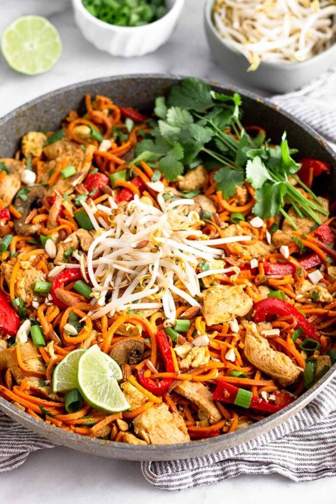 Large skillet filled with healthy chicken pad thai topped with bean sprouts, lime wedges, and fresh cilantro. Behind it is a bowl of bean sprouts, green onions, and a lime wedges.