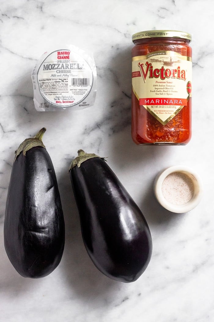 White counter top with a package of mozzarella cheese, a jar of marinara sauce, container of salt, and 2 eggplants on it.