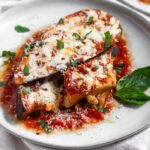 Healthy Instant Pot eggplant parmesan topped with fresh basil, red pepper flakes, and mozzarella cheese on a white that is sitting on a white linen.