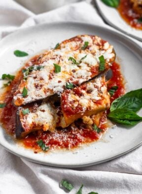 Healthy Instant Pot eggplant parmesan topped with fresh basil, red pepper flakes, and mozzarella cheese on a white that is sitting on a white linen.