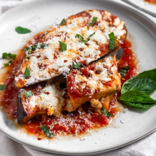 Healthy Instant Pot eggplant parmesan topped with fresh basil, red pepper flakes, and mozzarella cheese on a white that is sitting on a white linen.