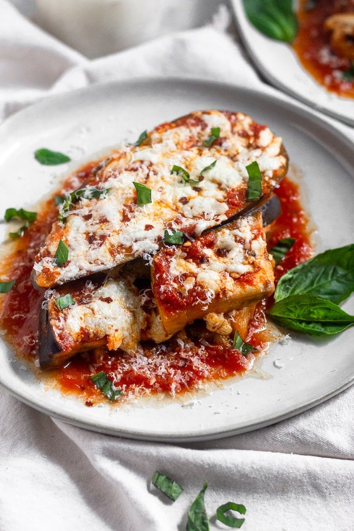 Healthy Instant Pot eggplant parmesan topped with fresh basil, red pepper flakes, and mozzarella cheese on a white that is sitting on a white linen.