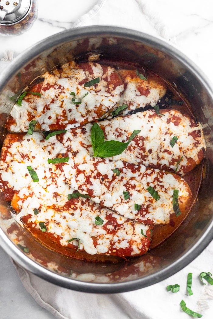 Healthy Instant Pot eggplant parmesan in the instant pot topped with fresh basil. It is sitting on a white linen with more basil and a red pepper flake shaker next to it.