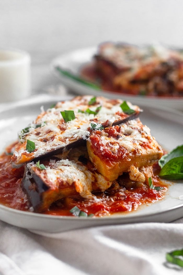Layers of instant pot eggplant parmesan topped with fresh basil sitting on a white plate. There is another plate behind it with more healthy eggplant parmesan on it.