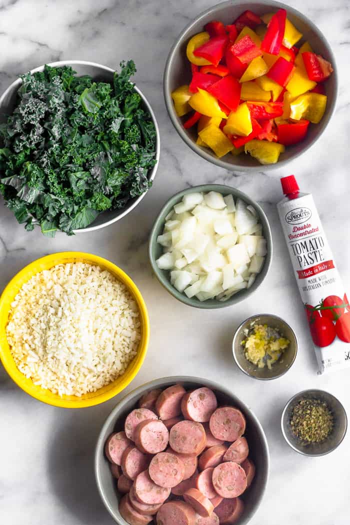White counter with the ingredients of sausage, pepper, and rice soup. There is kale, peppers, onion, tomato paste, garlic, spices, chicken sausage, and cauliflower rice.
