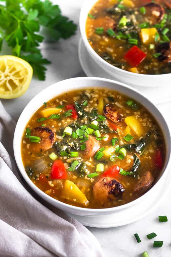 Two bowls of low carb sausage, pepper, and rice soup topped with green onions. A bunch of parsley and half a lemon is behind them and a tan linen is next to the front bowl.