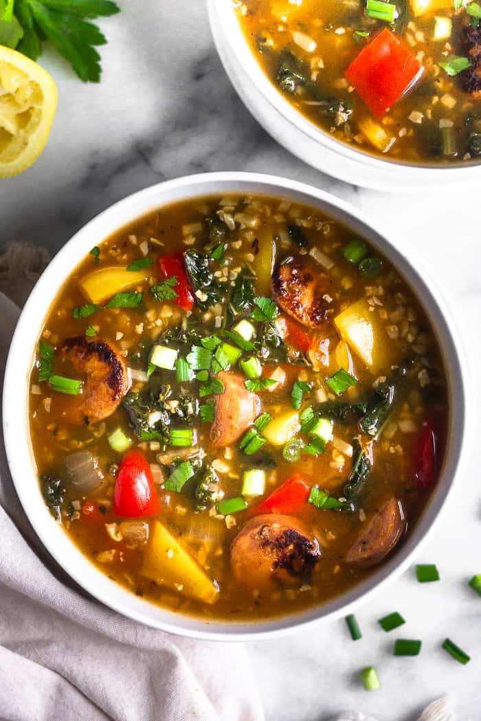 Overhead view of a white bowl filled with paleo sausage, pepper, and rice soup sprinkled with green onions. Around the bowl is more chopped green onion, half a lemon, some parley, a tan linen, and another bowl of soup.