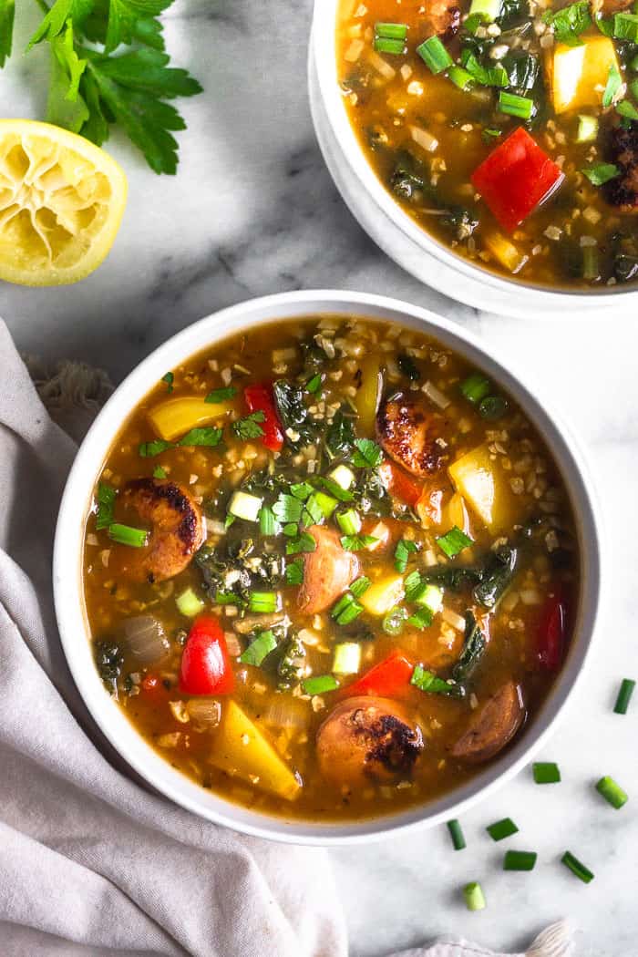 Overhead view of two white bowls filled with sausage, pepper, and rice soup sprinkled with green onions. Around the bowl is more chopped green onion, half a lemon, some parley, and a tan linen.