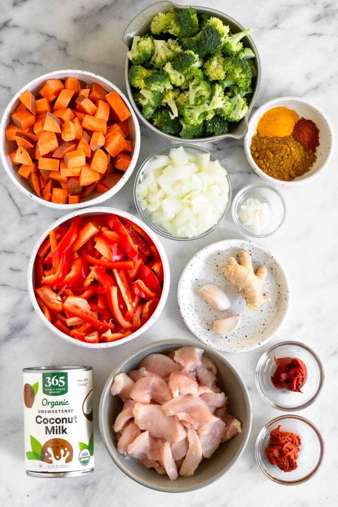 White counter with a bowl of broccoli florets, a bowl of spices, a bowl of coconut oil, a bowl of diced onions, a plate of 2 cloves of garlic and some fresh ginger, a bowl of curry paste, a bowl of tomato paste, a bowl of diced raw chicken, a can of coconut milk, a bowl of julienned red bell peppers, and a bowl of diced sweet potatoes.